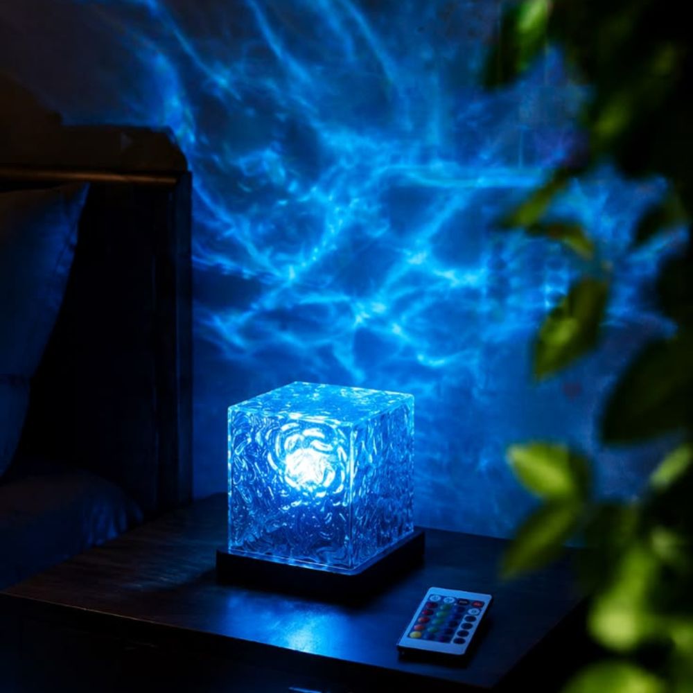 A glowing blue cube-shaped lamp sits on a dark bedside table, casting an underwater-like blue light pattern across the wall behind it. A small remote control lies nearby, with a bed and blurred green leaves visible in the room.